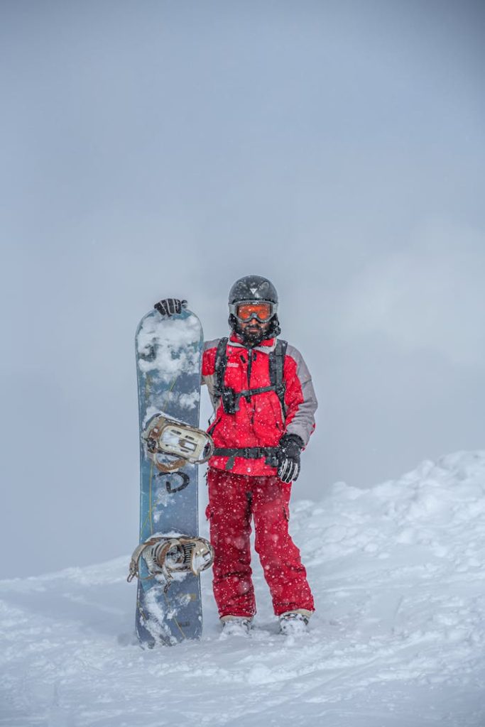 Snowboarder in red outfit stands with snowboard in snowy Gulmarg, capturing winter sports vibe.